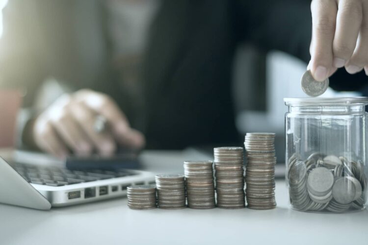 A person stacks coins next to a laptop and puts a coin into a glass jar, with four increasing stacks of coins arranged in front.