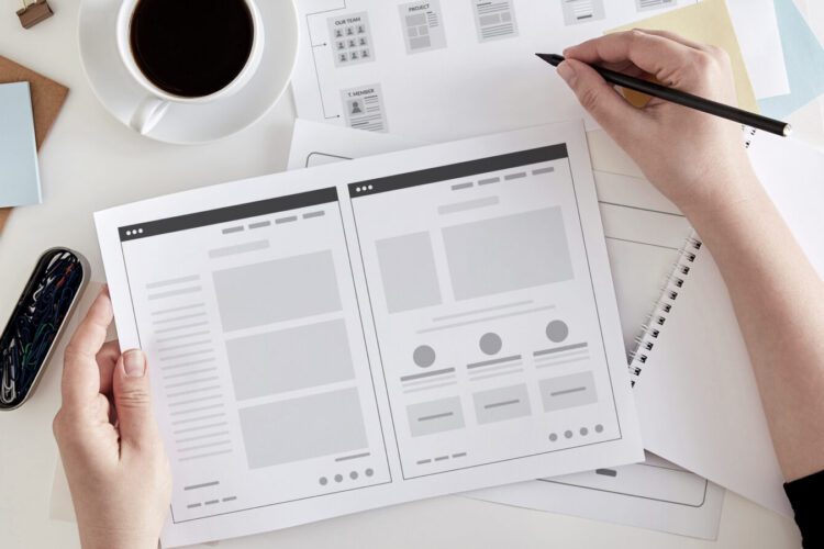 Person holding a website wireframe sketch at a desk with a cup of coffee, a notebook, pen, and various papers and sticky notes.