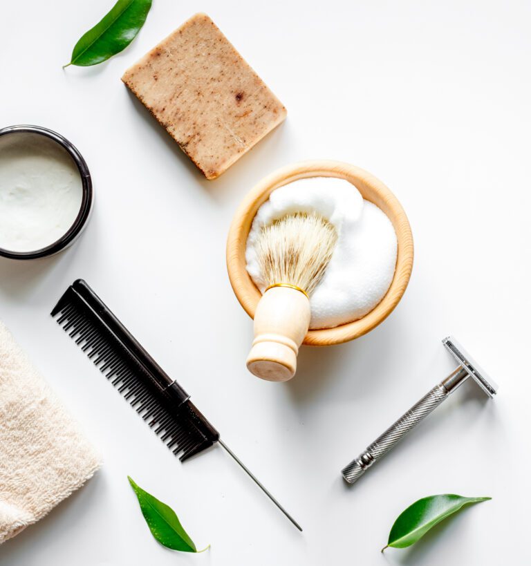 Flat lay of shaving items including a brush, shaving foam, safety razor, comb, soap bar, cream jar, towel, and green leaves on a white background.