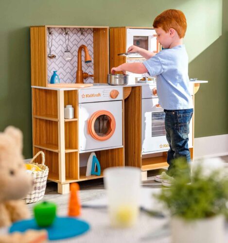 A young child plays with a wooden toy kitchen set, pretending to cook with a pot and spoon. Various kitchen accessories are visible, including a toy washing machine and sink.