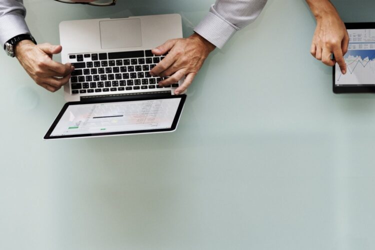 Two people working at a desk, one using a laptop and the other using a tablet displaying a graph.