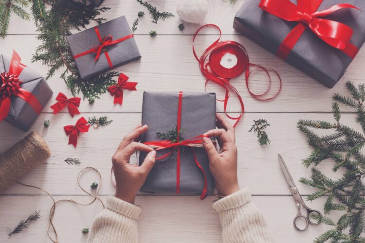 Hands tying a red ribbon around a gift box on a white wooden table surrounded by wrapped gifts, pine branches, ribbon, twine, and scissors.