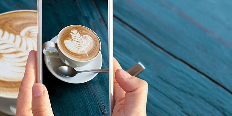 A person holds a smartphone to take a photo of a cup of coffee with latte art, on a saucer with a spoon, placed on a blue wooden table.