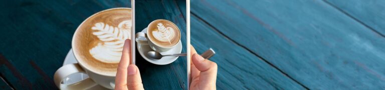 A person holds a smartphone to take a photo of a cup of coffee with latte art, on a saucer with a spoon, placed on a blue wooden table.
