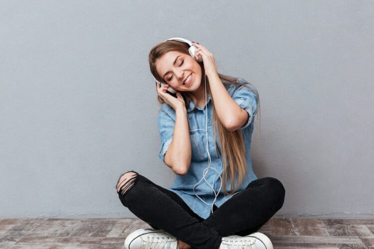 Young woman sitting cross-legged on the floor, wearing headphones, eyes closed, and smiling against a plain gray wall.