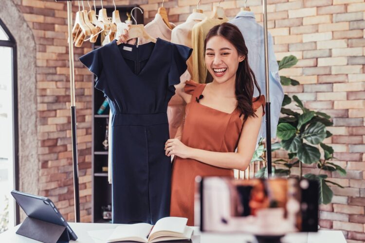 A woman stands in front of a clothing rack, holding a dress and smiling while recording a video with a smartphone, likely for an online fashion presentation.