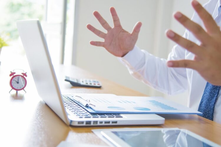 A person in business attire gestures with open hands at a desk with a laptop, charts, a calculator, alarm clock, and a tablet.