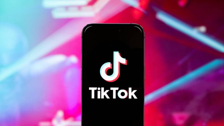 A smartphone displays the TikTok logo on its screen, with a blurred pink and purple background.