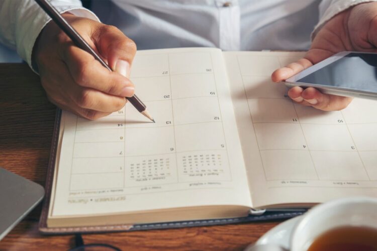 A person writes in a planner with a pencil while holding a smartphone, seated at a desk with a laptop and a cup of tea.