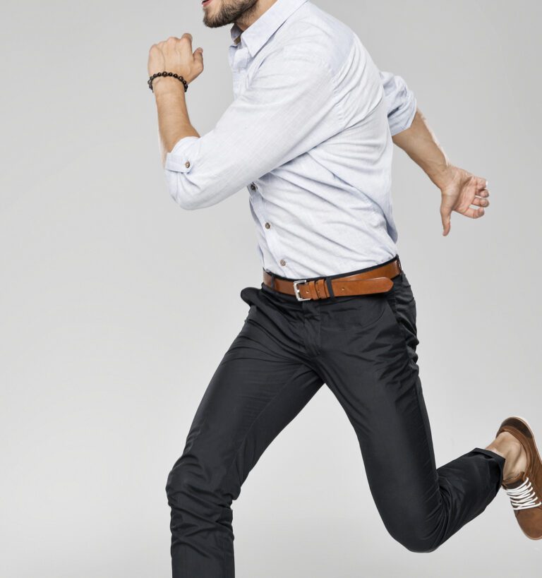 A man dressed in a light shirt, dark pants, and brown shoes is captured mid-run against a plain gray background.