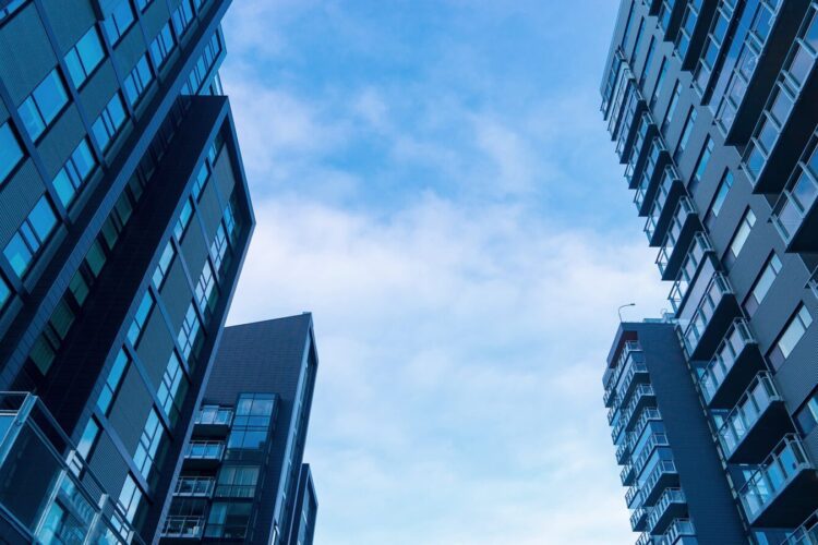 Tall modern apartment buildings with glass windows seen from below against a blue sky with light clouds.