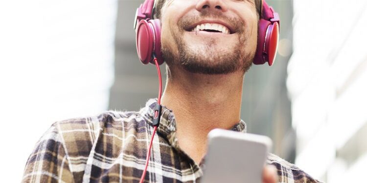 A man wearing a plaid shirt and red headphones smiles while holding a smartphone, standing outdoors with modern buildings in the background.