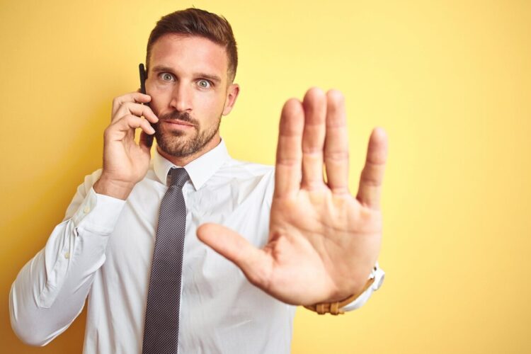 A man in a white shirt and tie holds a phone to his ear and raises his hand toward the camera in a stop gesture, standing against a yellow background.