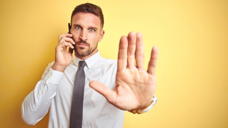 A man in a white shirt and tie holds a phone to his ear and raises his hand toward the camera in a stop gesture, standing against a yellow background.