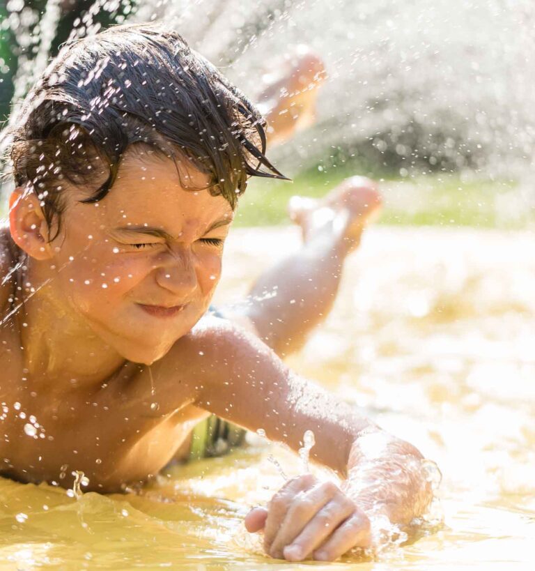 A boy slides on a yellow slip-and-slide in the sun, water splashing around him as he keeps his eyes closed and smiles.