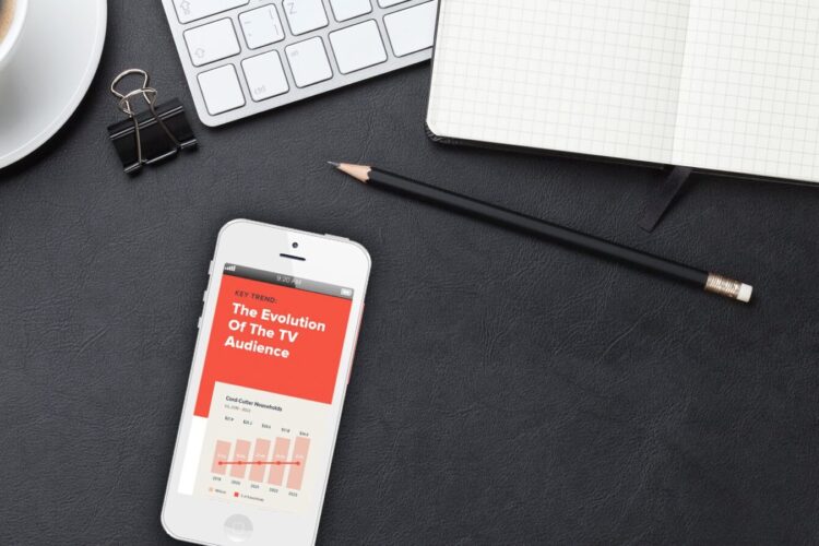 A smartphone displaying a TV audience report rests on a black desk with a keyboard, coffee, pencil, binder clip, and an open notebook.