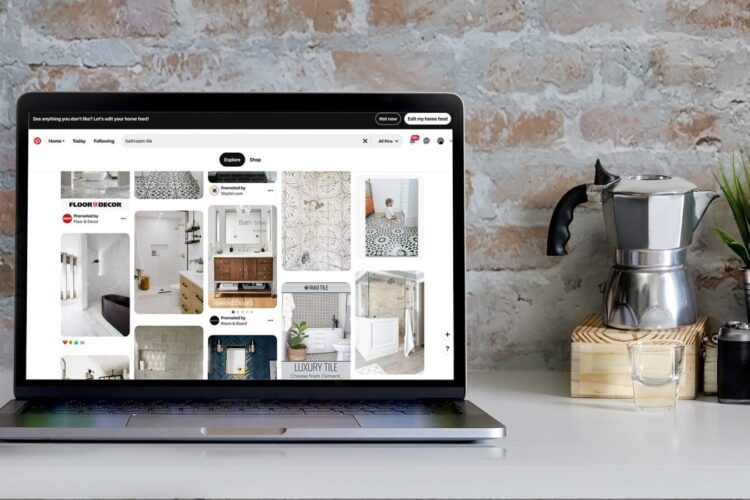 A laptop displaying a Pinterest page with home decor ideas sits on a table next to a moka pot, a camera, and a jar against a brick wall background.