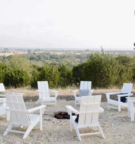 Six white Adirondack chairs are arranged in a circle around a fire pit on a gravel area, overlooking a landscape of trees and open fields under a cloudy sky.