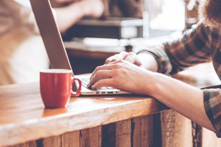 A person types on a laptop at a wooden counter next to a red mug, with another person blurred in the background.