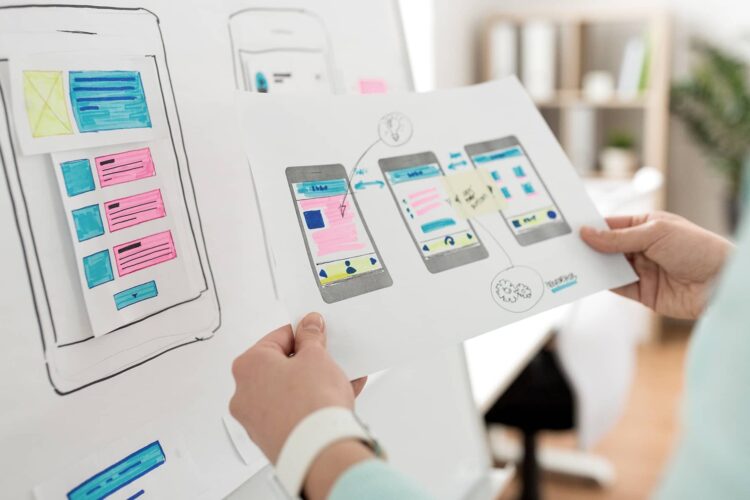 Person holding a paper with smartphone app wireframes and workflow sketches, with more wireframes displayed on a board in the background.