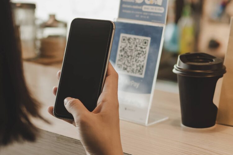 A person holds a smartphone near a counter with a QR code sign and a disposable coffee cup.