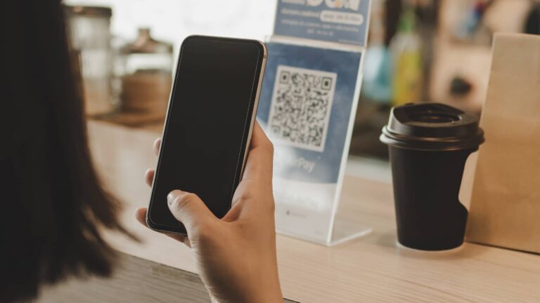 A person holds a smartphone near a counter with a QR code sign and a disposable coffee cup.