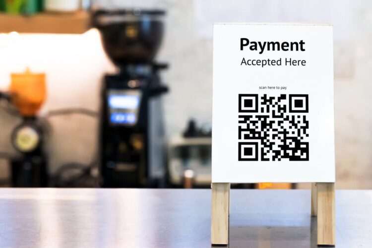 A payment sign with a QR code stands on a counter, indicating "Payment Accepted Here" and prompting users to scan to pay. Coffee shop equipment is blurred in the background.