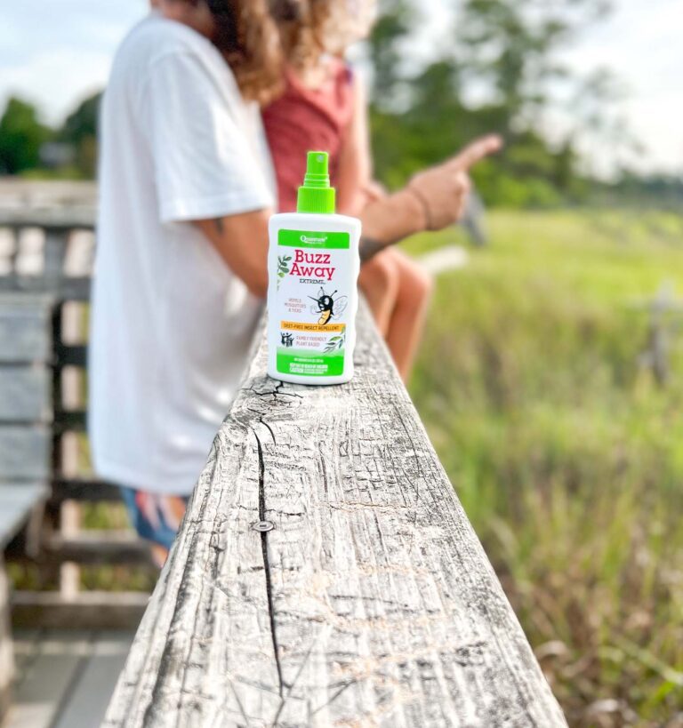 A bottle of Buzz Away insect repellent sits on a wooden railing, with two people sitting and talking in the blurred background near grassy wetlands.