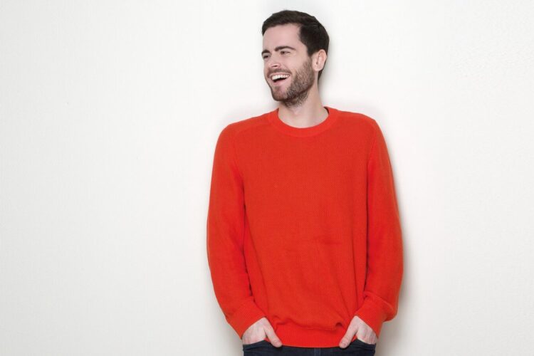 A man wearing a bright orange sweater stands against a white wall, smiling and looking to the side with his hands in his pockets.