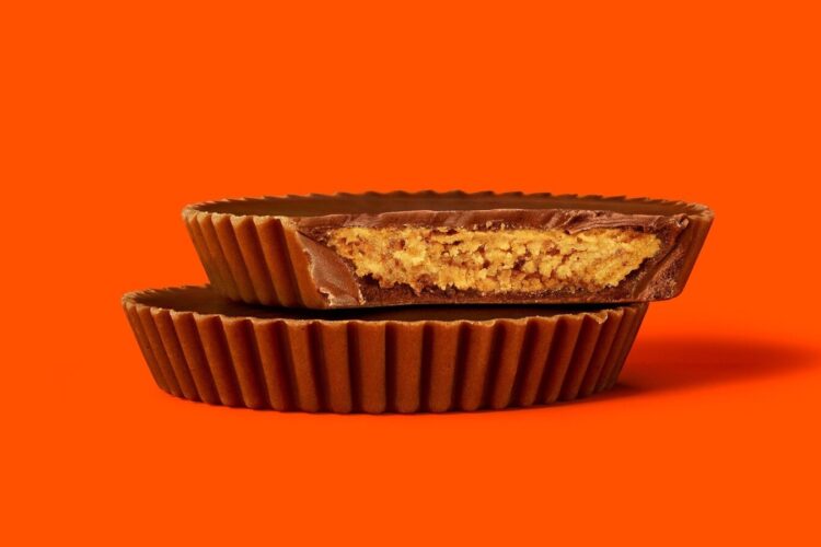 Two chocolate peanut butter cups are stacked, with the top one cut in half to show the peanut butter filling, set against an orange background.