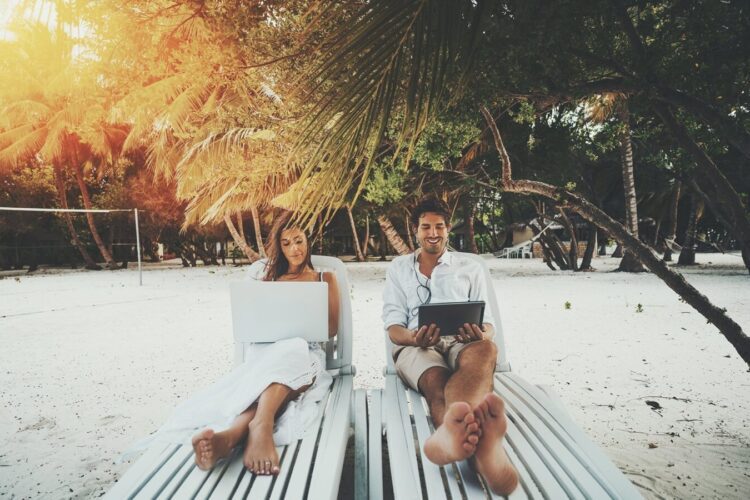 A man and woman relax on lounge chairs on a sandy beach under palm trees, both using laptops, with sunlight filtering through the trees.