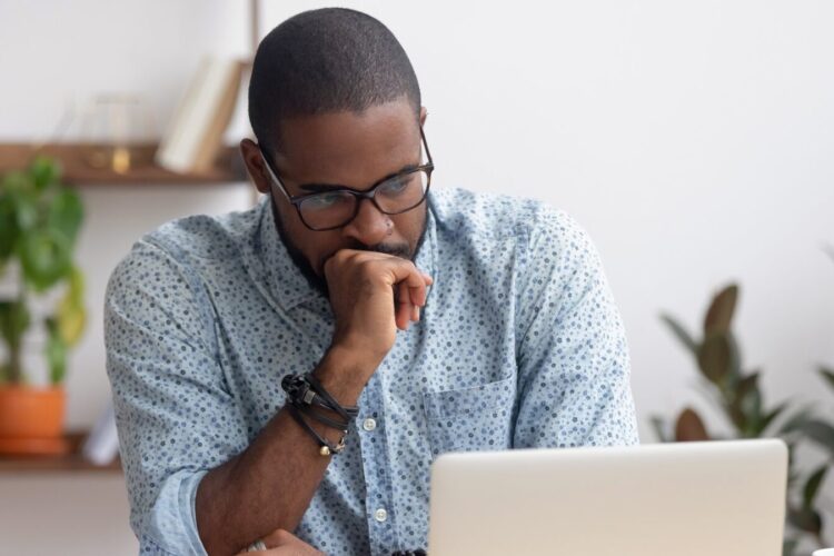 A man wearing glasses and a patterned shirt sits at a desk, looking thoughtfully at a laptop screen with his hand resting on his chin.