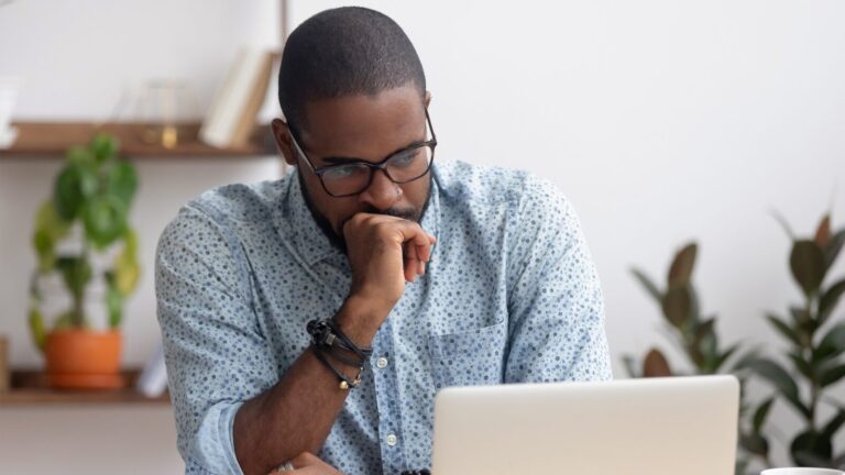 A man wearing glasses and a patterned shirt sits at a desk, looking thoughtfully at a laptop screen with his hand resting on his chin.