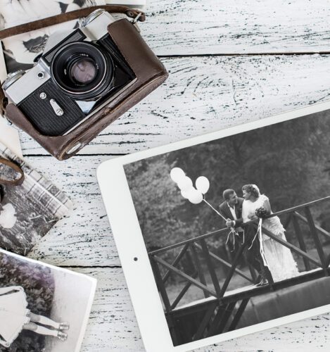 A tablet displays a black-and-white photo of a bride and groom on a bridge with balloons; a vintage camera and old photos rest on a white wooden surface nearby.
