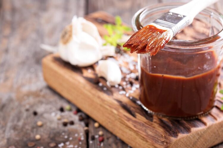A jar of barbecue sauce with a basting brush rests on a wooden board, alongside garlic cloves, peppercorns, and herbs.