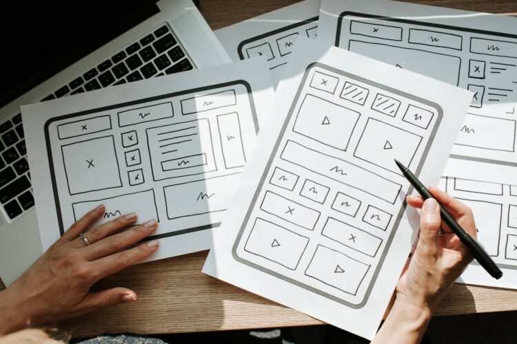 A person reviews website wireframe sketches on paper, with a laptop partially visible underneath the sketches on a wooden desk.
