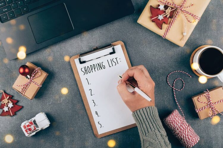 A hand writes on a shopping list on a clipboard beside a laptop, coffee cup, wrapped gifts, toy car, and string on a gray tabletop.
