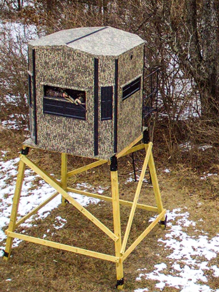 A camouflaged hunting blind on a wooden platform stands outdoors near leafless bushes and sparse snow on the ground.