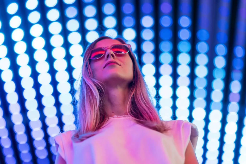 A person wearing red sunglasses and a white shirt stands in front of a background with rows of blue and white circular lights.