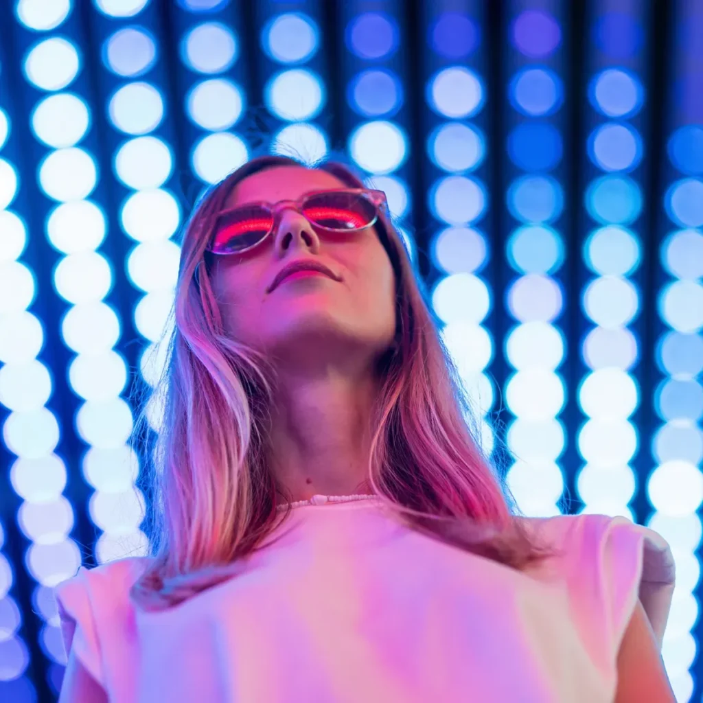 A woman wearing pink sunglasses and a white shirt stands in front of a wall of blue and white circular lights, looking upward.