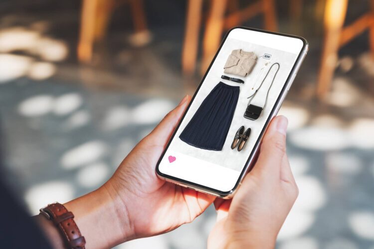 Person holding a smartphone displaying a photo of women's clothing and accessories, including a beige top, black skirt, shoes, bag, and necklace, arranged on a flat surface.