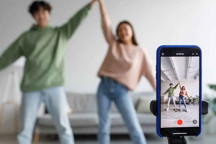 Two people stand indoors posing with raised arms, while a smartphone on a tripod records a video of them in front of a sofa.