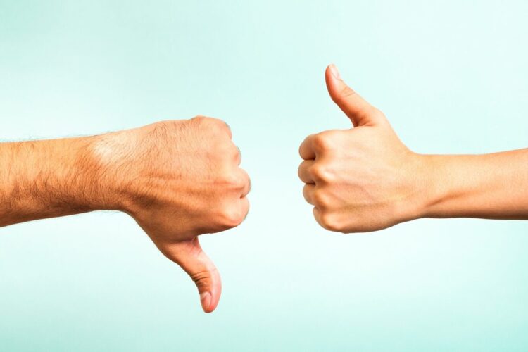 Two hands in front of a light blue background: one showing a thumbs down gesture, the other showing a thumbs up gesture.