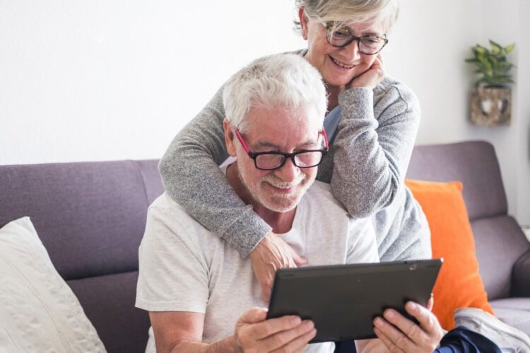 Two older adults sit on a couch, smiling while looking at a tablet device together in a well-lit living room.