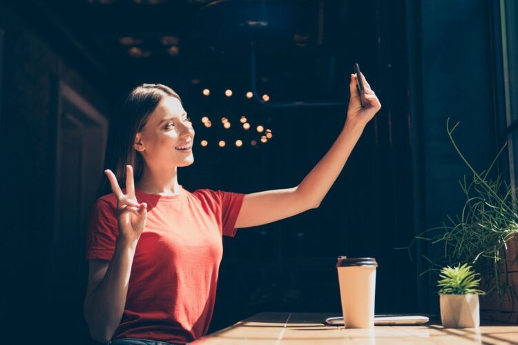 A woman in a red shirt sits at a table indoors, taking a selfie with her phone and making a peace sign. A coffee cup and small potted plants are on the table.