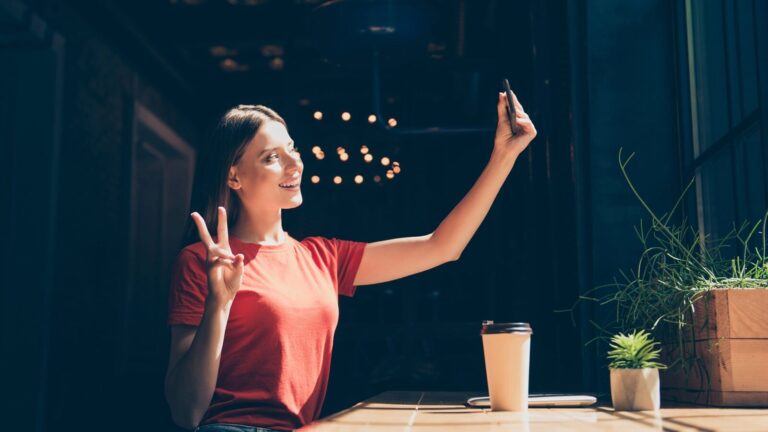 A woman in a red shirt sits at a table indoors, taking a selfie with her phone and making a peace sign. A coffee cup and small potted plants are on the table.