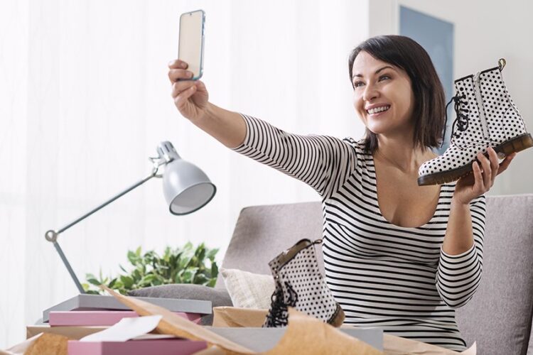 Woman sitting on a sofa holds up a polka dot boot and takes a selfie with her phone, surrounded by boxes and packing paper.