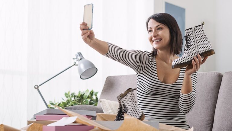Woman sitting on a sofa holds up a polka dot boot and takes a selfie with her phone, surrounded by boxes and packing paper.