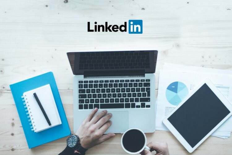 A person uses a laptop on a desk with LinkedIn logo above, surrounded by a notebook, documents, a tablet, and a coffee cup.