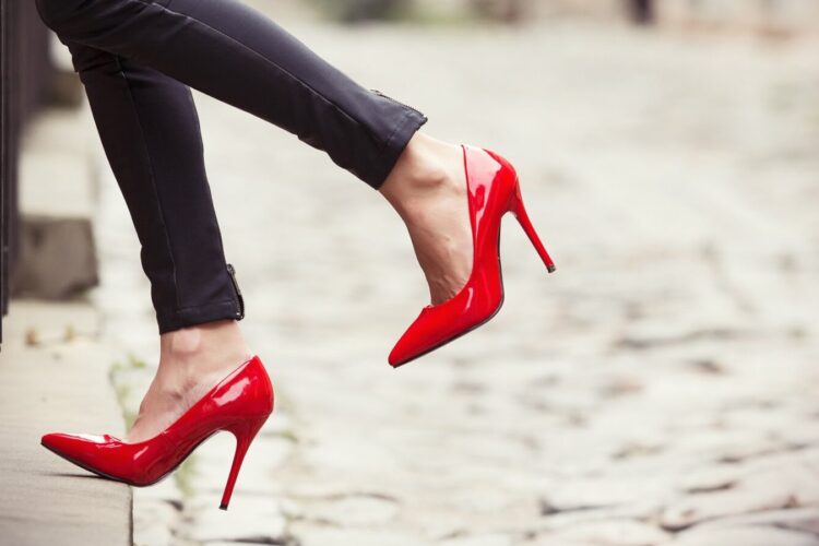 A person wearing black pants and red high-heeled shoes stands on a cobblestone street with one foot lifted.